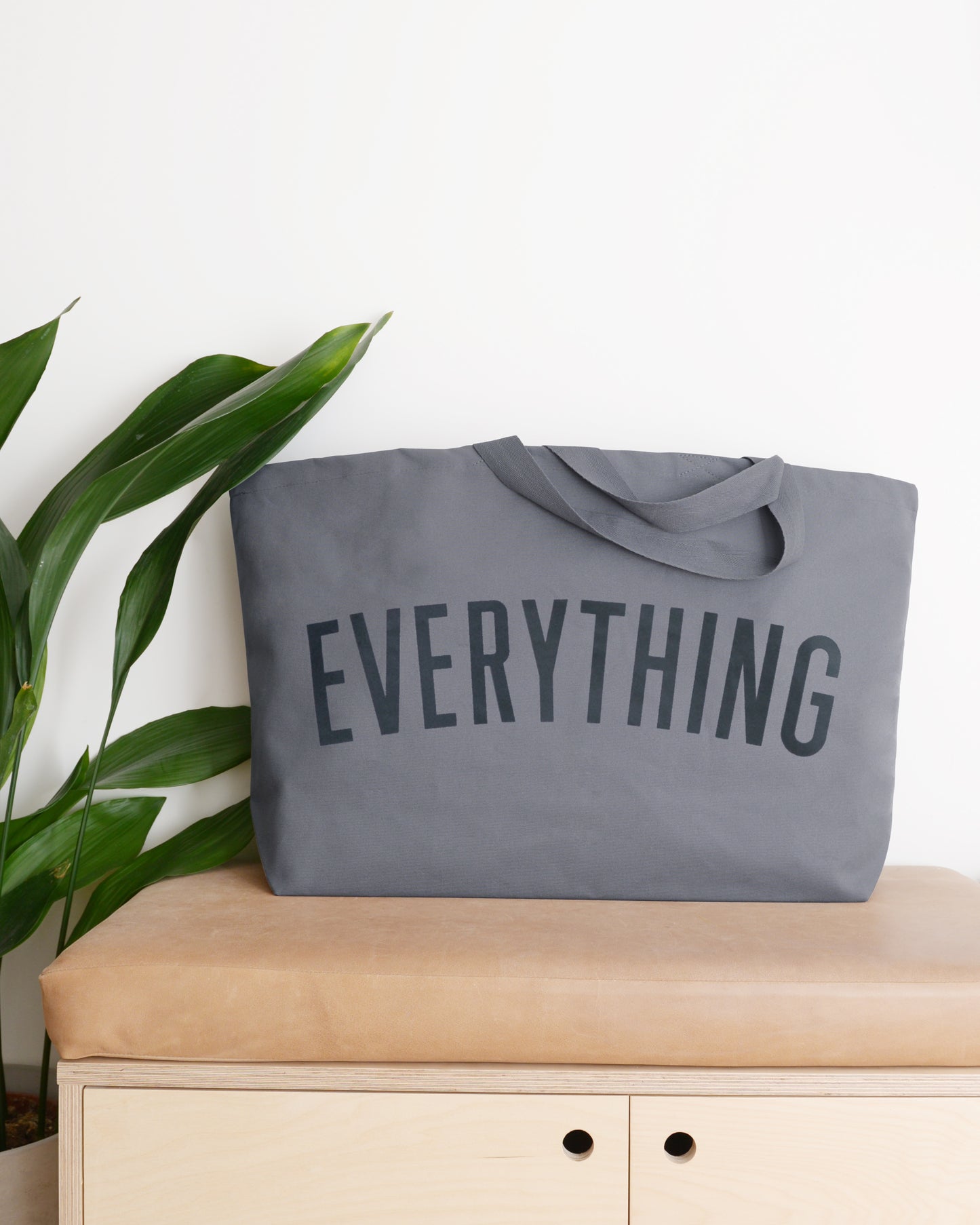 large grey tote bag screenprinted with the word everything in charcoal grey on a bench with a plant